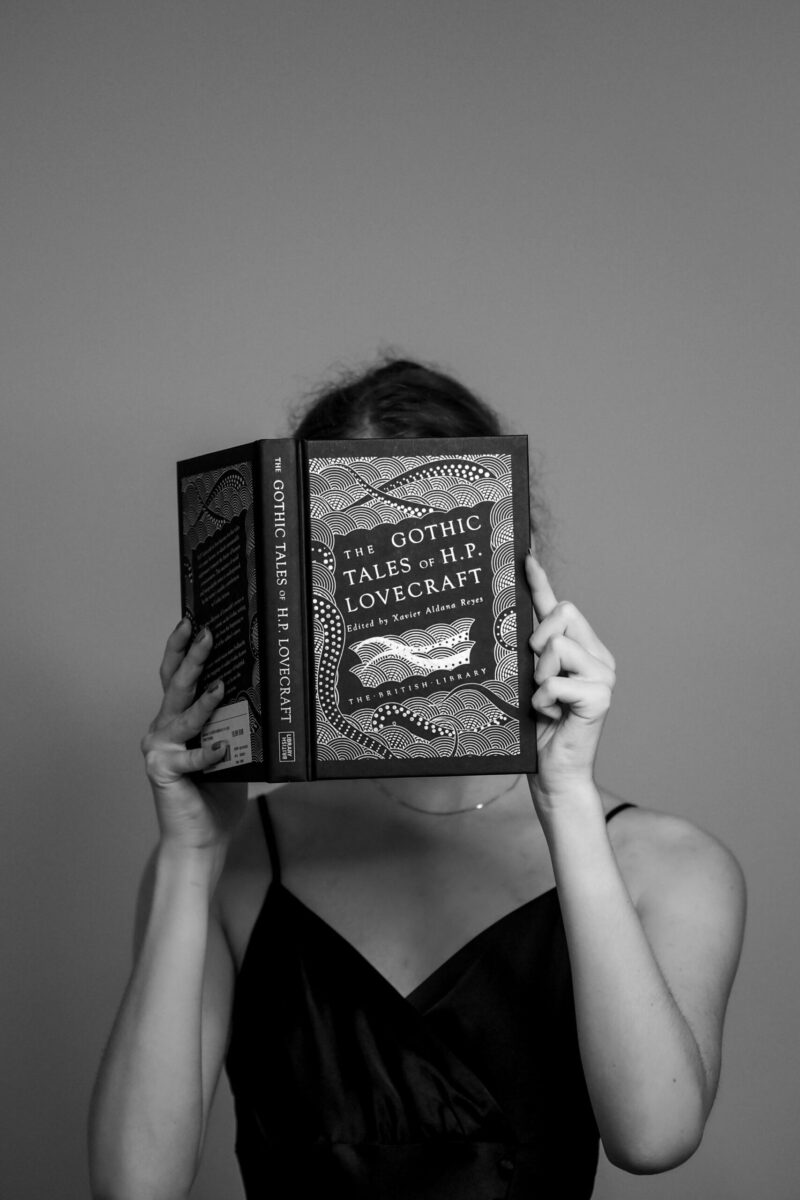 Black and white portrait of a woman reading 'The Gothic Tales of H.P. Lovecraft' in a studio setting.
